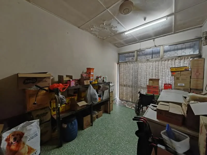 Room features: green terrazzo flooring, white painted walls, ceiling with visible damage, large window with sheer curtains, multiple cardboard boxes, shelves with various items, wheelchair, plastic bags, pet food bag, cluttered storage space.