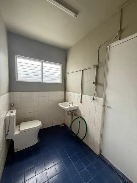 Bathroom features: white tiled walls, grey painted walls, blue ceramic tile flooring, one-piece toilet, wall-mounted sink, window with horizontal slats, exposed plumbing, door, light fixture.