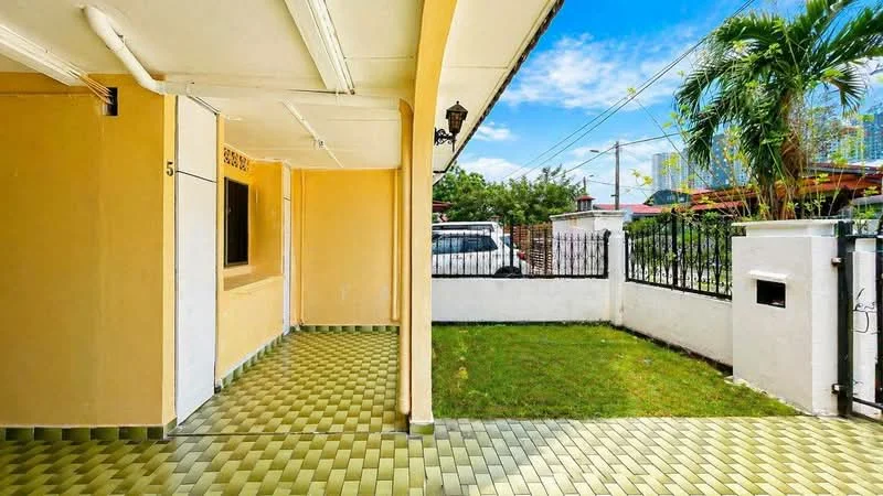 Exterior type: linked terrace house; features: yellow painted facade, covered porch area, green grass lawn, paved walkway, fenced perimeter, outdoor lighting fixture, window with security bars, car parked in driveway; style: modern.