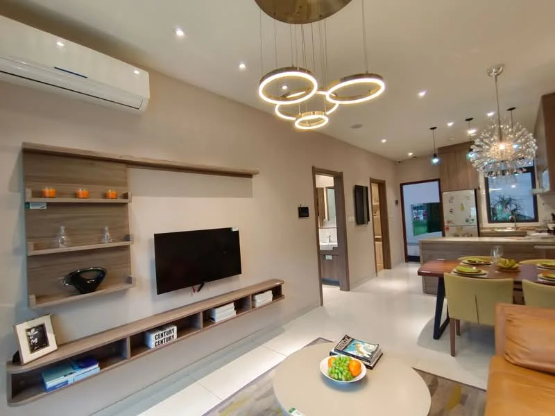 Living features: wall-mounted TV, wooden TV console, decorative wall shelf, 5-blade ceiling fan with integrated LED light, recessed downlights, large window with view, dining area with 4-seater table, open-plan layout, neutral painted walls, light-colored flooring, decorative chandelier, area rug with geometric pattern; style: modern; view: garden.