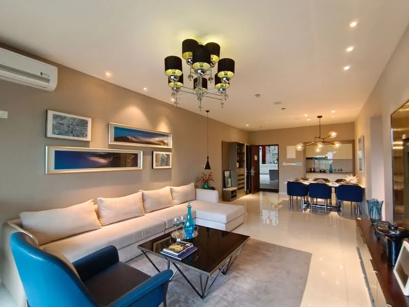 Living features: L-shape sectional sofa in cream, blue accent chair, glass coffee table, chandelier with multiple bulbs, recessed LED downlights, wall-mounted air conditioning unit, framed artwork on walls, decorative plant in ceramic pot, polished tile flooring, open layout to dining area; style: modern.