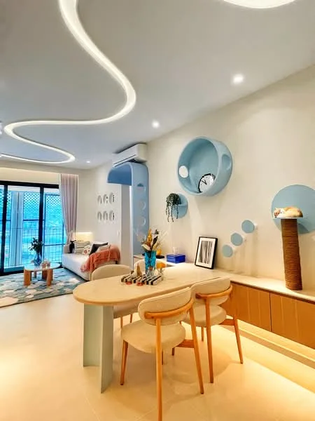 Dining features: round dining table with light blue base, 3 upholstered dining chairs in cream, decorative wall with circular blue accents, wall-mounted clock, light fixtures with curved design, floor-to-ceiling windows, sheer curtains, area rug with geometric pattern, wooden sideboard with storage, vase with flowers on the table, wall art framed in black; style: modern; view: garden. Living features: fabric sofa in light grey, throw blanket in coral, coffee table, decorative cushions, wall-mounted TV, area rug with geometric pattern; style: modern; view: garden.