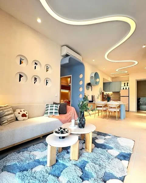 Living features: light blue accent wall, white painted walls, sofa with decorative cushions, round coffee table with white top, wooden legs on coffee table, area rug with blue and white patterns, recessed ceiling lights, air conditioning unit, dining area with wooden chairs, open kitchen with light cabinetry, decorative wall cutouts, greenery in vase; style: modern.