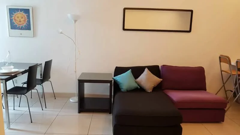 Living features: black fabric sectional sofa with purple accent, glass coffee table, floor lamp with white shade, framed artwork with sun design, dining table with four black chairs, tile flooring, mirror on wall, decorative cushions in blue and purple; style: modern.