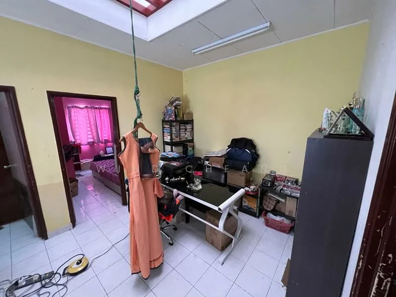 Room features: yellow painted walls, white tiled flooring, ceiling light fixture, window with sheer curtains, hanging dress on a hook, black office chair, glass-top table, boxes and storage items, wooden cabinet, bed visible in background.
