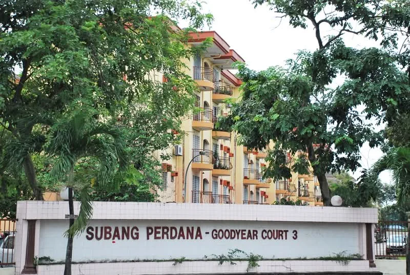 Exterior type: low-rise apartment block; features: cream painted facade, recessed balconies, multiple air conditioning units visible, landscaped greenery in foreground, signage with 'Subang Perdana - Goodyear Court 3', surrounded by trees, two-storey building, brick wall at the base; style: modern.