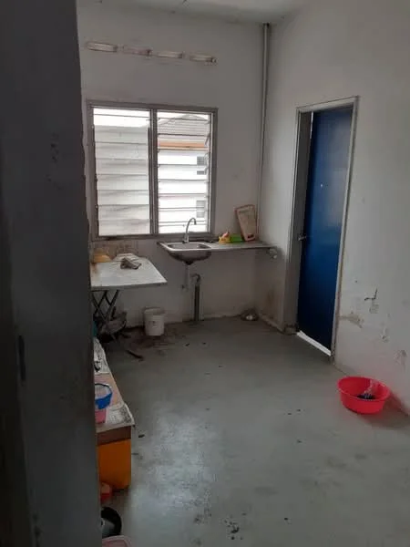 Room features: white painted walls, concrete flooring, single-bowl stainless-steel sink, window with horizontal slats, blue door, table with folding legs, bucket, pink basin, visible plumbing, minimal decor; style: minimalist.