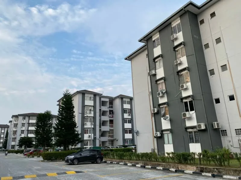 Exterior type: low-rise apartment block; features: grey and white painted facade, multiple air conditioning units visible, landscaped garden area in front, manicured lawn, parking lot with marked spaces, surrounded by trees, two-storey building, windows with grilles; style: modern.