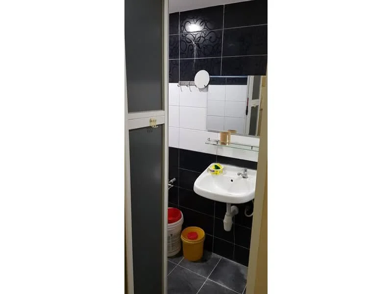 Bathroom features: white ceramic sink, chrome basin mixer faucet, mirror above sink, black tiled walls, white tiled walls, grey floor tiles, shower area, toilet, plastic storage bucket, plastic waste bin.