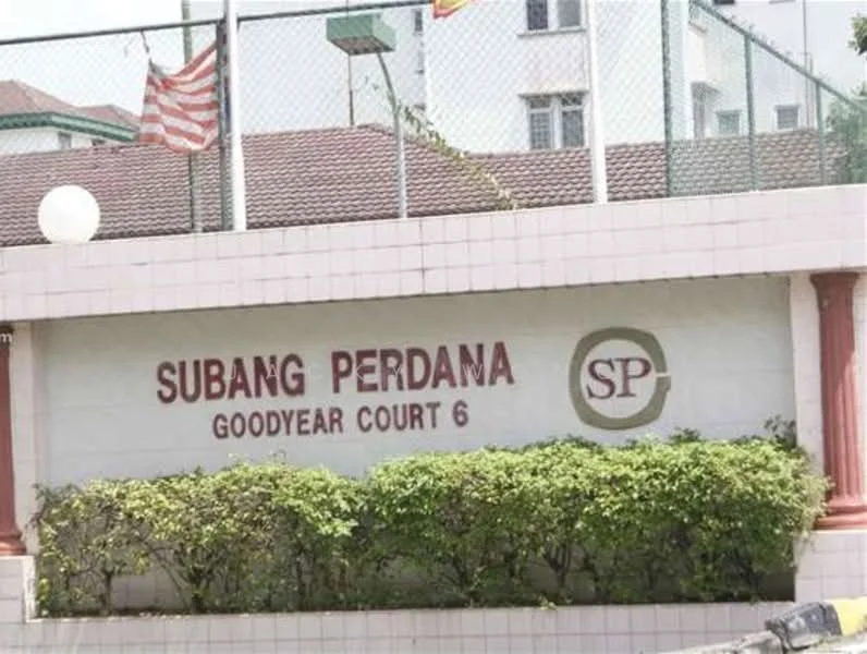 Exterior features: signage with 'SUBANG PERDANA GOODYEAR COURT 6', landscaped greenery, brick wall, fence surrounding the area, flag poles, residential buildings in background; style: modern.