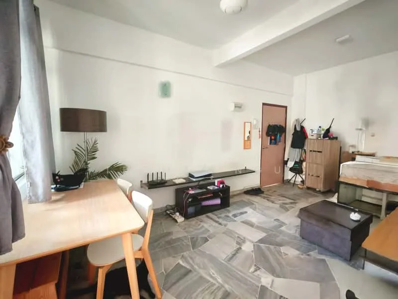 Room features: grey stone flooring, white painted walls, ceiling light fixture, large window with sheer curtains, wooden desk with chair, wooden storage cabinet, black storage ottoman, small potted plant, clothes hanging on wall hooks, bed with bedding visible; style: modern.