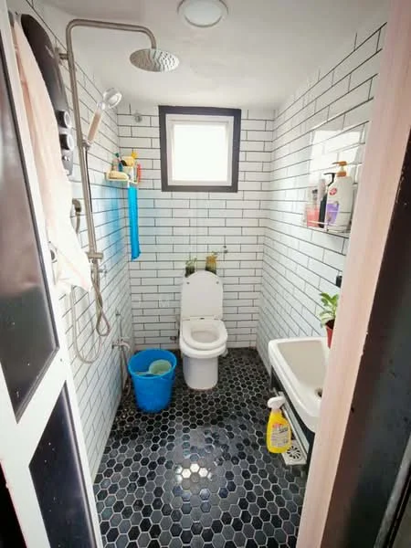 Bathroom features: white tiled walls with black grout, black pebble tile flooring, walk-in shower with glass enclosure, rain shower head, handheld shower, toilet, small sink, window for natural light, shower caddy with toiletries, cleaning supplies, potted plant; style: modern.