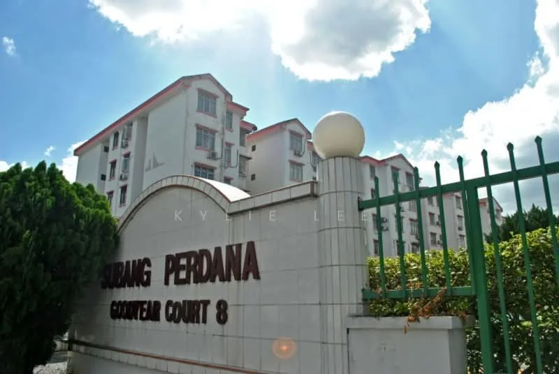 Exterior type: low-rise apartment block; features: white painted facade, red tiled roof, green fencing, landscaped garden with shrubs, signage with 'Subang Perdana' and 'Court 8', multiple storeys, surrounded by greenery, clear blue sky; style: modern.