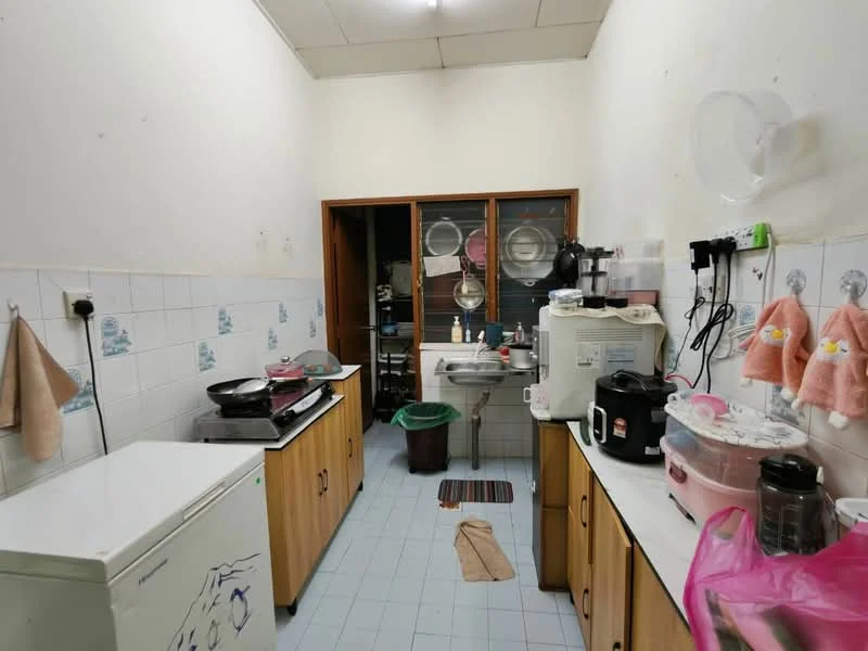 Kitchen features: white tiled walls, light blue tiled flooring, wood cabinetry with laminate finish, double-bowl stainless-steel sink, chrome faucet, freestanding gas stove, white refrigerator, microwave, rice cooker, kettle, countertop with space for appliances, hanging towels, window with metal grille, garbage bin; style: functional.