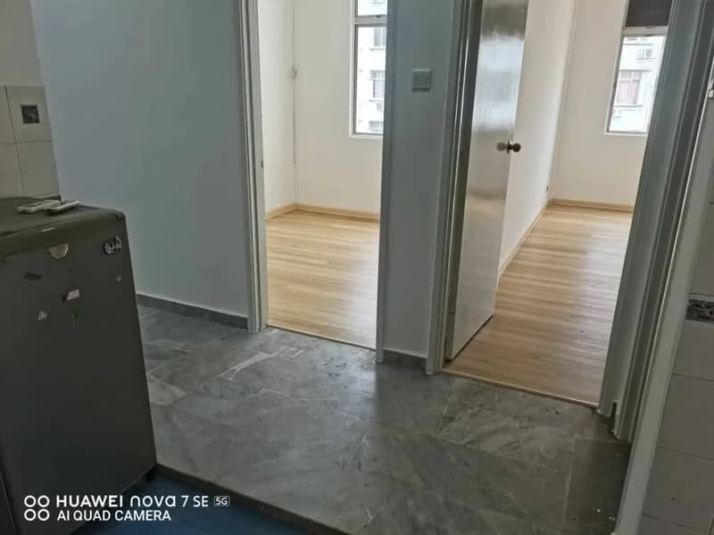 Room features: grey marble flooring, light wood laminate flooring, white painted walls, doorway leading to another room, window with natural light, kitchen area visible with fridge, neutral color scheme; style: modern.