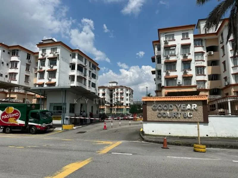 Exterior type: low-rise apartment block; features: white painted facade, orange accent trim, multiple storeys, main entrance gate, guard house at entrance, surrounded by greenery, paved driveway, parking area, signage at entrance; style: modern.