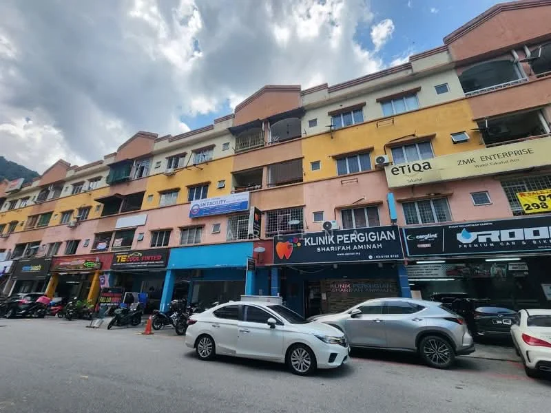 Exterior type: shophouse; features: multi-storey building, mixed-use commercial and residential, colorful facade with orange and yellow paint, balconies on upper floors, signage for various businesses, parking spaces in front, motorcycles parked along the street, cars parked along the street, urban setting with nearby hills; style: modern.