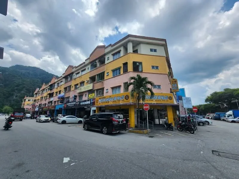 Exterior type: shophouse; features: 3-storey building, yellow and pink painted facade, balconies on upper floors, ground floor retail space, signage visible on facade, parking spaces in front, surrounded by vehicles, mountain backdrop, urban street view; style: modern.