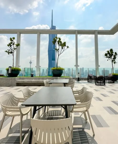 Exterior type: rooftop terrace; features: outdoor dining table, white plastic chairs, planters with greenery, city skyline view, glass railing, paved flooring, sunny weather, surrounding urban landscape; style: modern.