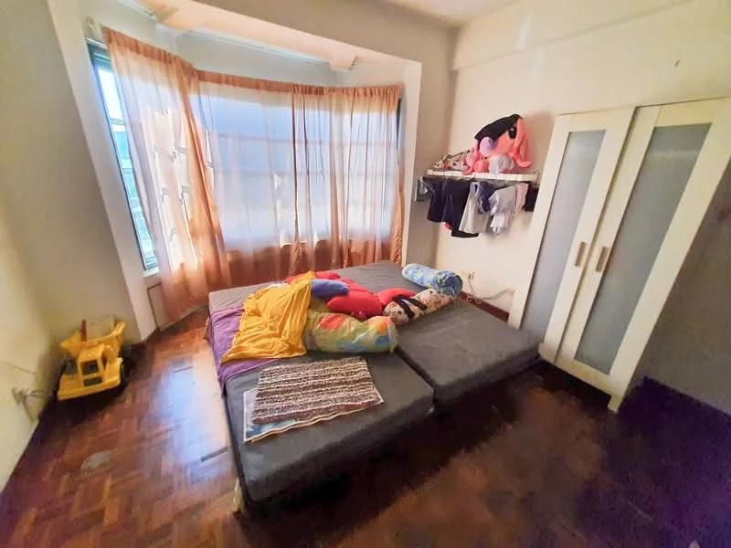 Bedroom features: wooden flooring, large windows with sheer curtains, bedding with colorful pillows and blankets, built-in wardrobe with sliding doors, wall-mounted shelf with clothing, corner layout with bay window, decorative wall with light color, natural light from windows.
