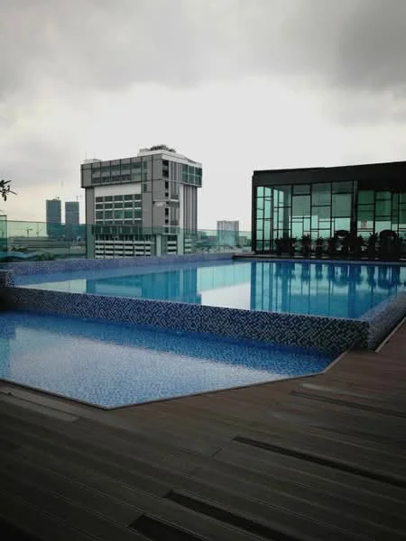 Exterior type: swimming pool deck; features: infinity pool, swimming pool with mosaic tile edging, wooden deck flooring, surrounding city skyline, glass facade building in background, outdoor seating area with chairs, cloudy sky; style: modern.