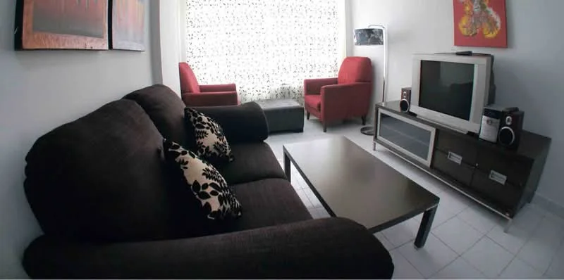 Living features: 3-seater fabric sofa in dark brown, decorative cushions with floral pattern, coffee table with metal legs and glass top, two red armchairs, TV console with storage drawers, flat-screen TV, speakers on either side of the TV, framed artwork on walls, white sheer curtains, white walls, tile flooring; style: modern.