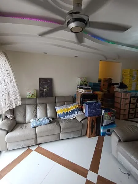 Living features: 3-seater fabric sofa in beige, decorative throw pillow with geometric pattern, ceiling fan with integrated LED light, light-colored walls, tile flooring with brown accent pattern, large window with sheer curtains, framed artwork on wall, various boxes and storage items stacked, area rug, wall-mounted light fixture; style: modern.