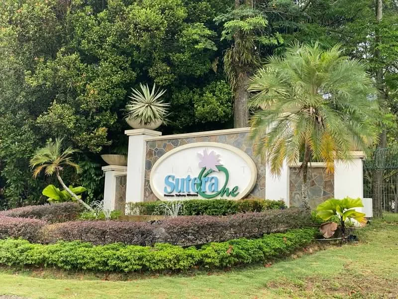Exterior type: main entrance gate; features: landscaped garden with shrubs, stone cladding on entrance wall, decorative water feature, palm trees, tropical landscaping, signage with 'Sutera One', manicured lawn, flower beds; style: tropical-modern.