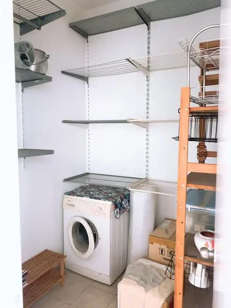 Laundry features: front-load washing machine, metal shelving units, wooden storage rack, tile flooring, white painted walls, open storage space, baskets, laundry supplies.