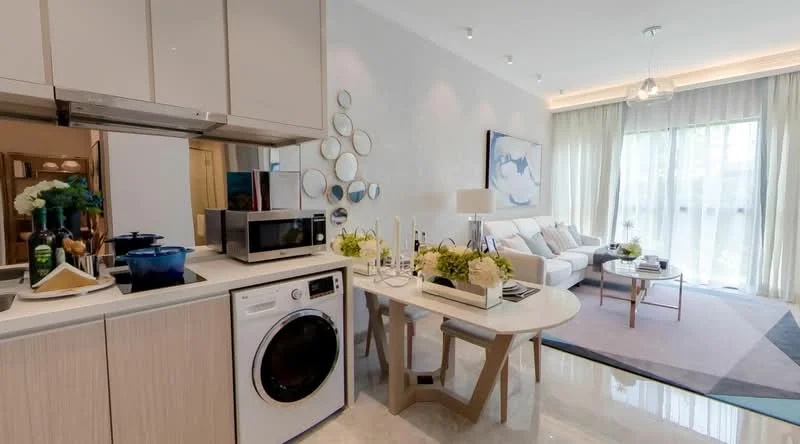 Kitchen features: white cabinetry, built-in microwave, stainless-steel fridge, front-load washing machine, countertop with decorative items, kitchen utensils, blue cooking pot, open shelving; style: modern. Living features: light grey sofa, coffee table, decorative pillows, area rug with geometric pattern, wall-mounted artwork, floor-to-ceiling windows, sheer curtains, decorative plants; style: modern.