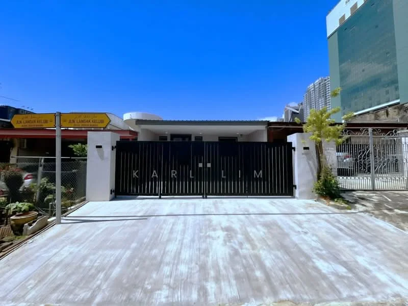 Exterior type: detached bungalow; features: black metal gate, white painted facade, flat roof, concrete driveway, landscaped garden with plants, surrounded by high-rise buildings, signage on the left, fenced perimeter; style: modern.