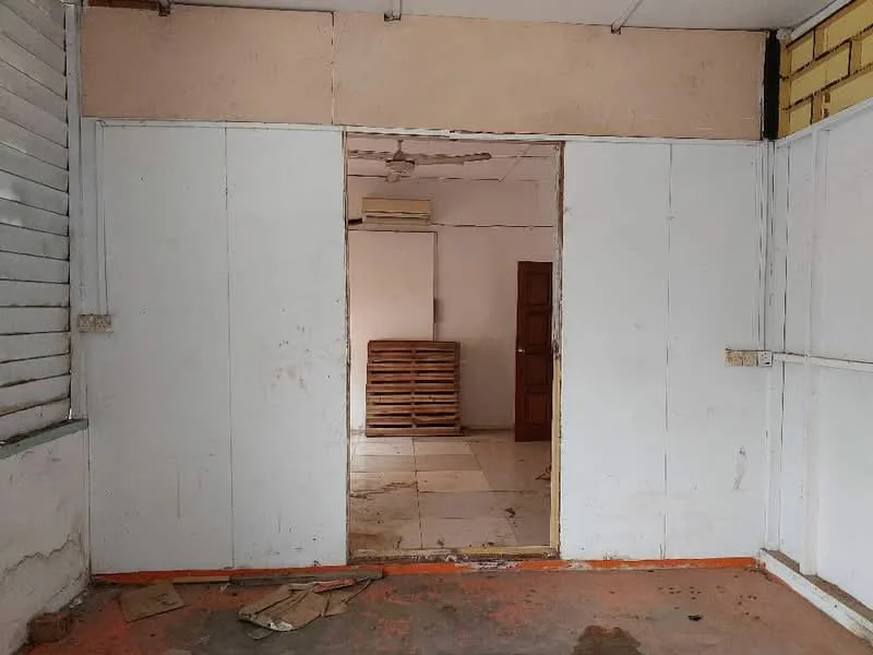Room features: white painted walls, exposed concrete flooring, ceiling fan, window with wooden slats, doorway leading to another room, stack of wooden pallets in background, tile flooring visible in adjacent room, orange baseboard.