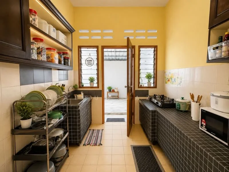 Kitchen features: yellow painted walls, black tiled countertops, open shelving with glass jars, stainless-steel microwave, stainless-steel gas stove, metal dish rack, ceramic plates and bowls, kitchen utensils, floor tiles, two windows with sheer curtains, door leading to outdoor area, rug at entrance; style: modern; view: courtyard.