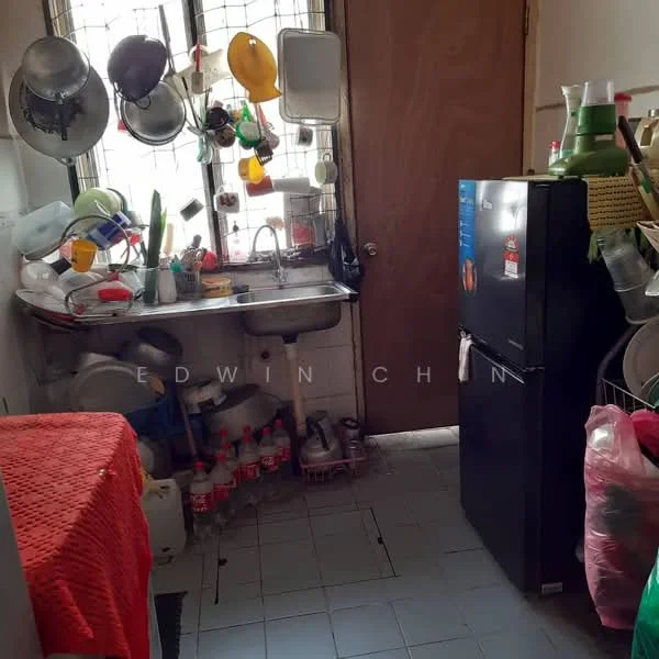 Kitchen features: single-bowl stainless-steel sink, chrome pull-down faucet, black fridge, wooden cabinet, plastic containers, metal pots, hanging utensils, red tablecloth, tile flooring, window with metal grille, shelves with various kitchen items, bottled drinks on the floor.