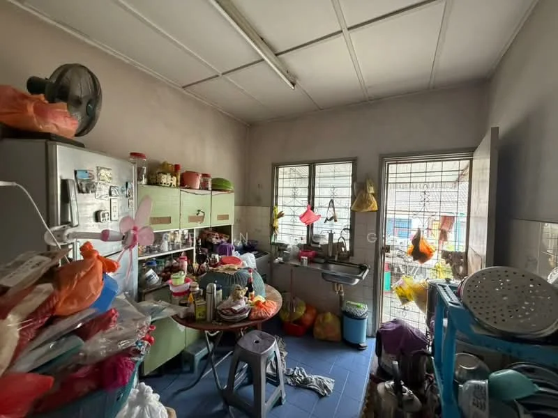 Kitchen features: grey painted walls, blue tile flooring, metal ceiling fan, double-bowl stainless-steel sink, chrome pull-down faucet, green cabinetry, refrigerator, countertop with clutter, multiple plastic bags and containers, table with assorted items, two windows with metal grilles.