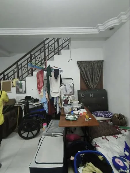 Room features: white painted walls, ceramic tile flooring, staircase with wooden railing, sofa, wooden table, various items on table, clothes hanging on railing, bed with bedding, laundry baskets, curtains, wall-mounted artwork, cluttered space.
