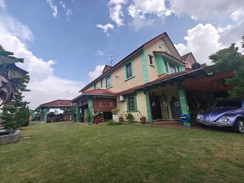 Exterior type: detached bungalow; features: green painted facade, red tiled roof, two-storey structure, manicured lawn, paved walkway, car porch with awning, outdoor seating area, mature trees, garden area, visible antenna on roof, classic car parked, surrounded by greenery; style: traditional.