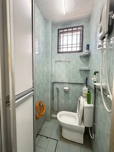 Bathroom features: light green tiled walls, light green tiled flooring, one-piece toilet, shower with handheld showerhead, wall-mounted shower mixer, mirror above sink, shelf for toiletries, window for natural light, water heater, toilet paper holder, soap dispenser, cleaning supplies, plastic storage container.