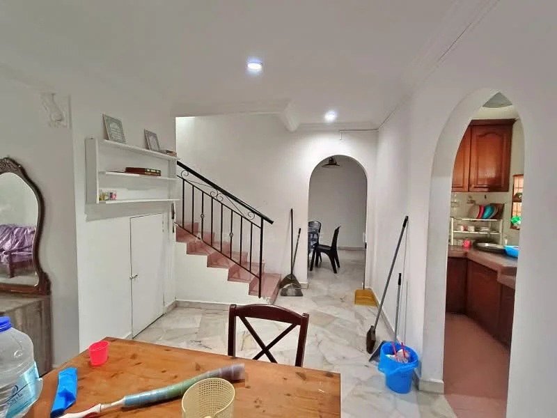 Room features: white painted walls, marble flooring, staircase with metal railing, arched doorways, built-in shelving unit, wooden dining table, plastic chairs, cleaning tools visible, kitchen area visible in background, recessed ceiling lights; style: modern.