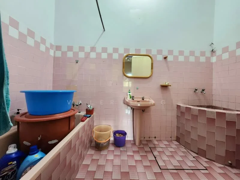 Bathroom features: pink tiled walls, pink tiled flooring, built-in bathtub, sink with pedestal, mirror above sink, shower fixtures, blue plastic basin, plastic waste bin, cleaning supplies, towel hanging on wall.