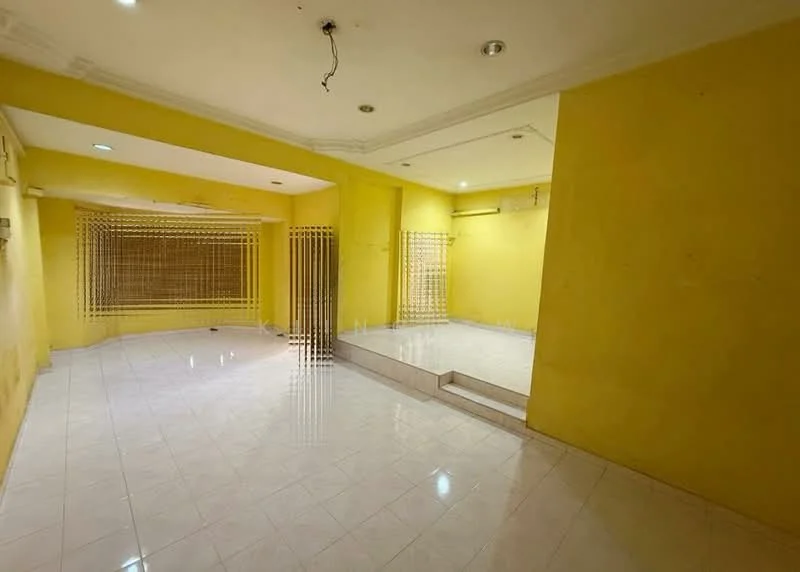 Room features: yellow painted walls, glossy white tile flooring, recessed ceiling lights, step-up platform area, metal security grille, large window with grille, open layout, no visible furniture.