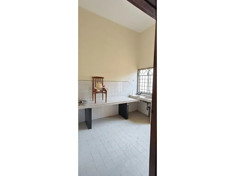 Kitchen features: white painted walls, light grey tiled flooring, single-bowl stainless-steel sink, wall-mounted faucet, countertop with storage underneath, window with metal grille, empty space for appliances, wooden chair.