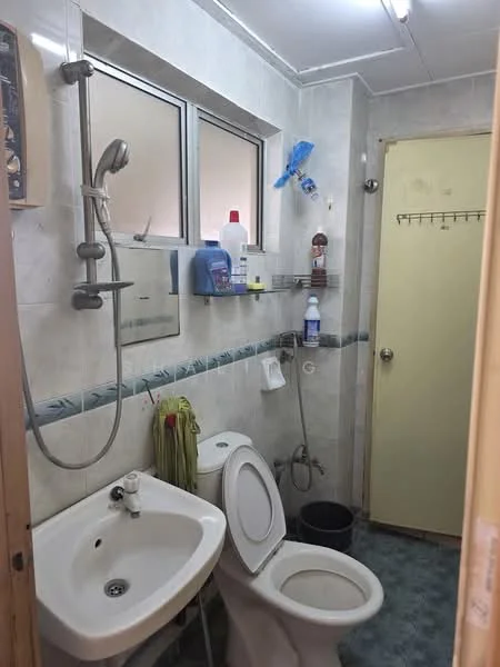 Bathroom features: white ceramic toilet, single-sink vanity with chrome faucet, mirror above sink, shower with handheld showerhead, wall-mounted soap dispenser, tiled walls with light grey tiles, blue vinyl flooring, window for natural light, shelf with cleaning products, green towel hanging, toilet paper holder, plastic waste bin.