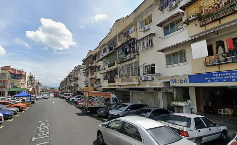 Exterior type: low-rise apartment block; features: multi-storey building, balconies with metal railings, various window types, ground-level parking, street-side shops, visible signage, cars parked along the street, urban setting with nearby mountains, cloudy sky; style: traditional.