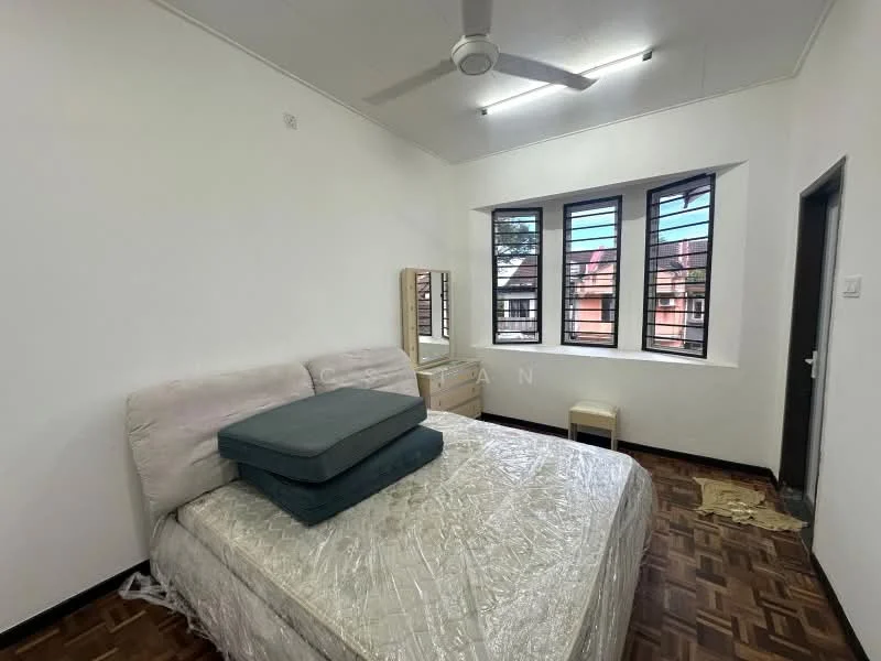 Bedroom features: queen-size bed, mattress with protective cover, wooden flooring, white painted walls, ceiling fan, three-pane window with black grills, dressing mirror, small stool, natural light from window.