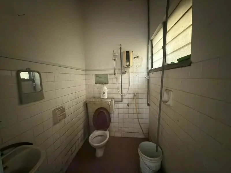 Bathroom features: white tiled walls, brown tiled flooring, one-piece toilet, shower with handheld showerhead, wall-mounted shower valve, mirror above sink, window with horizontal slats, bucket on the floor.