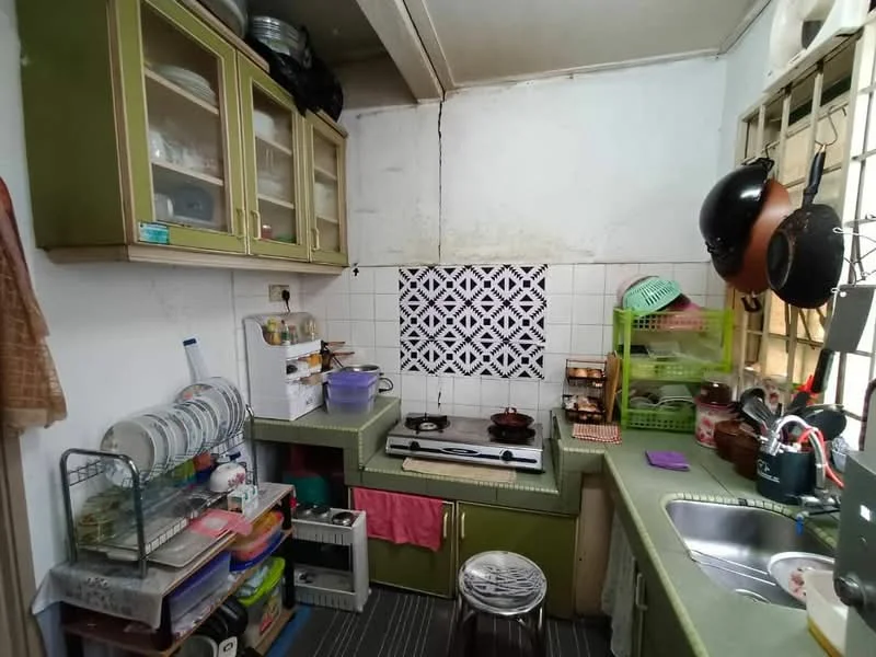 Kitchen features: green cabinetry, white tiled backsplash with geometric pattern, stainless-steel sink, single-bowl sink, gas stove, countertop with green surface, open shelving, dish rack, various kitchen utensils, window with metal grille, flooring with grey stripes, plastic storage containers.