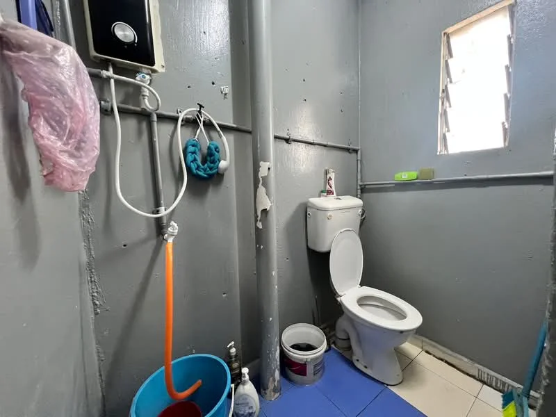 Bathroom features: grey painted walls, white toilet, shower with water heater, orange shower hose, blue bucket, white ceramic floor tiles, window with metal bars, plumbing pipe, cleaning supplies, toilet paper holder.