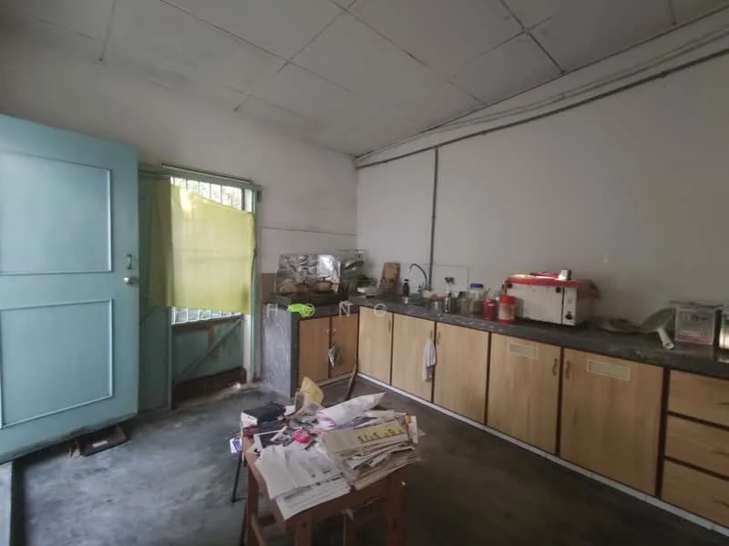 Kitchen features: green wooden door, yellow curtain, grey countertop, wooden cabinetry with handles, stainless-steel sink, freestanding white appliance, various kitchen utensils, concrete flooring, window with metal bars.