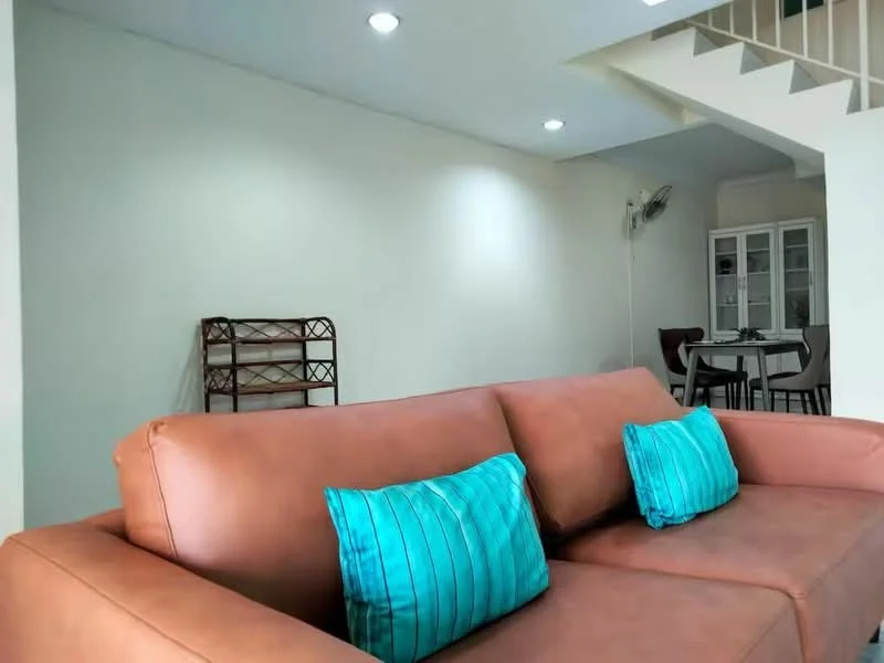 Living features: brown leather sofa, turquoise striped cushions, white painted walls, recessed downlights, ceiling fan, wooden shelving unit, dining table with chairs in background, open layout; style: modern.